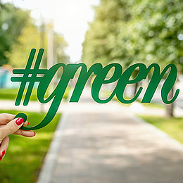 Eine weiße Hand hält Schriftzug "#green" vor einem Park im Hintergrund ©Artem Beliaikin / unsplash.com