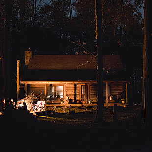 Eine Gruppe Menschen sitzt am Lagerfeuer vor einer Blockhütte ©Teddy Kelley / unsplash.com
