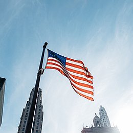 US-Flagge vor einem blauen Himmel und Wolkenkratzern ©Steven Abraham / unsplash.com