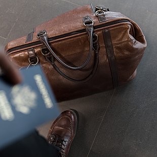 braune Ledertasche zu Füßen einer Person mit Reisepass in der Hand ©nappy / pexels.com