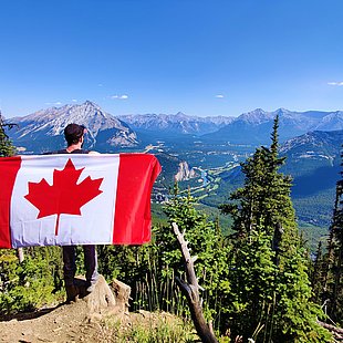 Junger Mann mit kanadischer Flagge blickt auf weite Berglandschaft ©Maxime Doré / unsplash.com